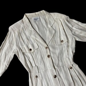 May include: A cream-colored, vertically striped dress with a collared neckline, long sleeves, and button-down front. The dress features two chest pockets and decorative buttons. The label reads "Bill Blass Studio".