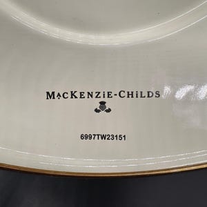 Op de afbeelding: Een cr&egrave;mekleurig keramieken bord met een goudkleurige rand. Het midden bevat de zwarte tekst MACKENZIE-CHILDS boven een klein distelpictogram en de alfanumerieke code 6997TW23151, gedrukt in zwarte inkt op het gladde, glanzende oppervlak.