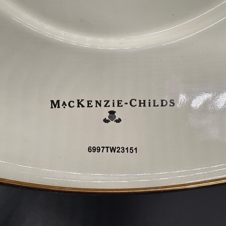 Op de afbeelding: Een cr&egrave;mekleurig keramieken bord met een goudkleurige rand. Het midden bevat de zwarte tekst MACKENZIE-CHILDS boven een klein distelpictogram en de alfanumerieke code 6997TW23151, gedrukt in zwarte inkt op het gladde, glanzende oppervlak.