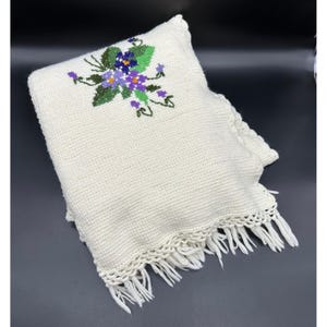 May include: Cream-colored crocheted blanket with a floral embroidery design. The embroidery features purple and blue flowers with green leaves. The blanket has a scalloped edge with fringe.