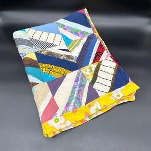 May include: A colorful patchwork quilt with a variety of fabric patterns and colors, including blue, pink, yellow, and white. The quilt has a yellow floral border and is folded. The quilt is a handmade item.