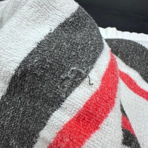 Op de afbeelding: Close-up van een witte handdoek met dikke grijze en rode strepen. De handdoek heeft een getextureerd oppervlak en een klein los draadje. De handdoek is opgevouwen en toont het strepenpatroon.