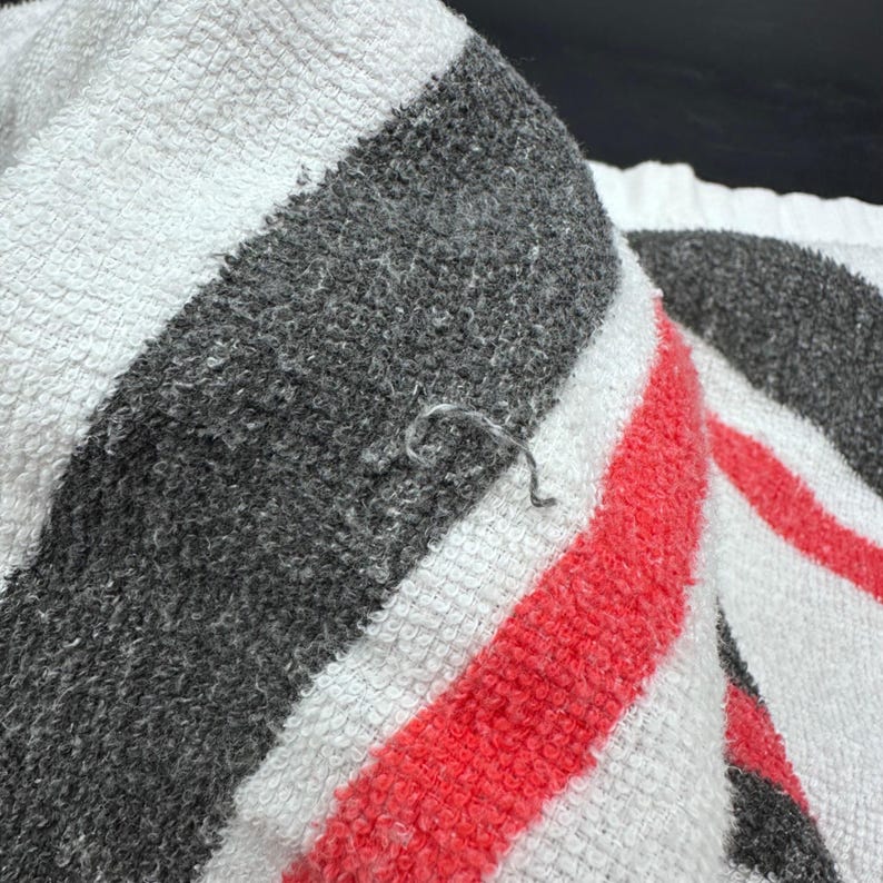 Op de afbeelding: Close-up van een witte handdoek met dikke grijze en rode strepen. De handdoek heeft een getextureerd oppervlak en een klein los draadje. De handdoek is opgevouwen en toont het strepenpatroon.