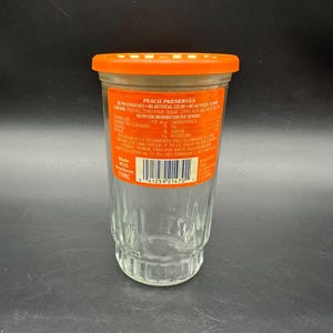 May include: A clear glass jar with an orange lid, labeled "PEACH PRESERVES." The label includes nutrition information and the text "Made With Southern Pride." The jar is empty, with a ridged design on the lower portion.