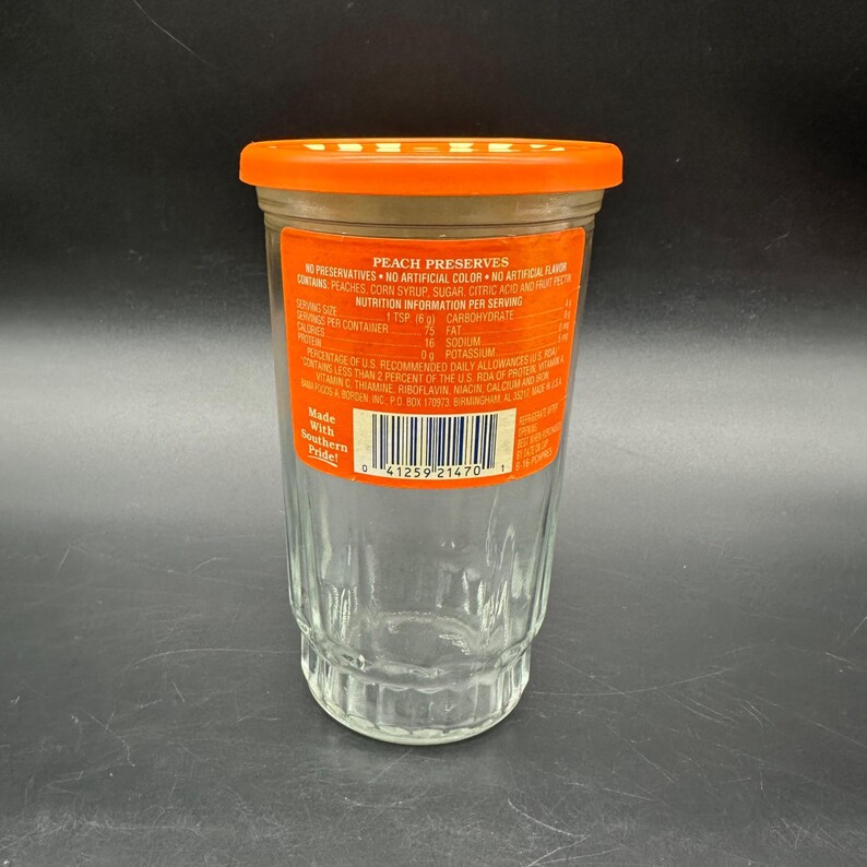 May include: A clear glass jar with an orange lid, labeled "PEACH PRESERVES." The label includes nutrition information and the text "Made With Southern Pride." The jar is empty, with a ridged design on the lower portion.