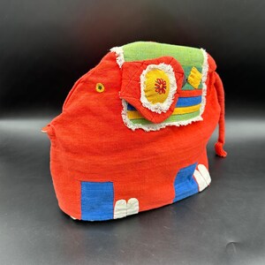 May include: A red fabric elephant-shaped tea cozy featuring a green, yellow, and blue patterned ear with white fringe. The design includes blue rectangular leg accents, white fabric toes, a small yellow eye, and a thin red tail on the back.