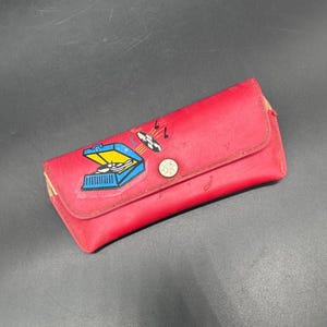 May include: A red vinyl glasses case with a button closure. The case features a colorful graphic of a record player with musical notes. The case is rectangular and has a slightly worn appearance.