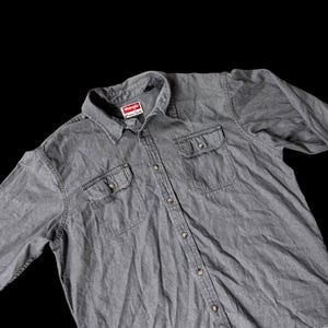 May include: A gray Wrangler button-down shirt with two chest pockets and a collar. The shirt has a faded appearance and a red and white Wrangler label on the collar.