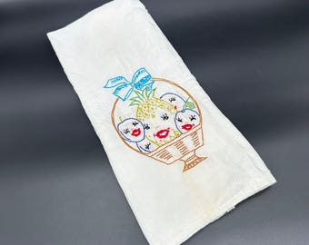 May include: White kitchen towel with embroidered fruit basket. The basket contains a pineapple and three plums with faces. The plums have red lips and eyelashes. The basket has a blue bow.