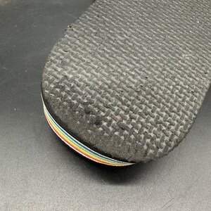 May include: Close-up of the sole of a black flip-flop. The sole has a textured pattern and a rainbow-colored strap. The flip-flop is likely made of rubber or a similar material.