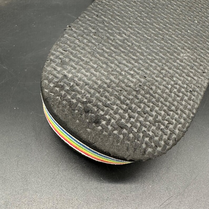May include: Close-up of the sole of a black flip-flop. The sole has a textured pattern and a rainbow-colored strap. The flip-flop is likely made of rubber or a similar material.