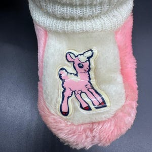 Peut inclure: Une pantoufle en peluche rose et blanche avec une application d'agneau de dessin anim&eacute;. La pantoufle a un revers blanc et un ext&eacute;rieur rose et duveteux. L'agneau est rose avec un contour bleu et des accents rouges.