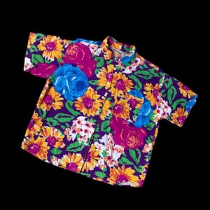 May include: Short-sleeved button-down shirt with a vibrant floral pattern. The shirt features large sunflowers, roses, and other flowers in shades of yellow, pink, blue, and white against a purple background. The shirt has a classic collar and a pocket.