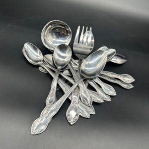 May include: A selection of vintage silverware, including spoons, forks, and a ladle, arranged on a dark surface. The silverware has ornate floral detailing on the handles and a shiny, reflective surface.