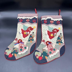 以下が含まれることがあります： パッチワークデザインのクリスマスストッキング2つ。各ストッキングには、赤い天使、緑のクリスマスツリー、ハート型のアクセントが施されています。ストッキングは赤と白のチェック柄のトップと赤い吊り下げループが付いています。