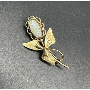 May include: A gold-toned brooch in the shape of a flower. The brooch features an oval-shaped, iridescent stone set within a scalloped frame. The flower design includes stylized leaves and a bow.