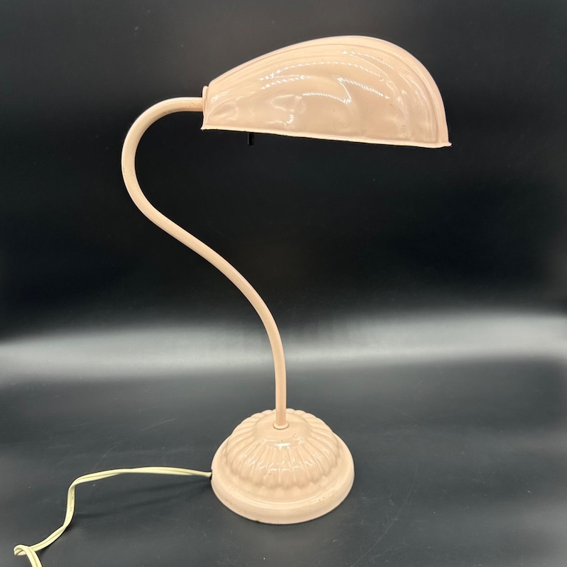 K&ouml;nnte beinhalten: Eine Vintage-Schreibtischlampe in einem weichen, ged&auml;mpften Rosa. Die Lampe hat einen gebogenen Schwanenhalsarm und einen gew&ouml;lbten Schirm. Der Sockel hat ein dekoratives, geriffeltes Design. Das Kabel ist cremefarben.