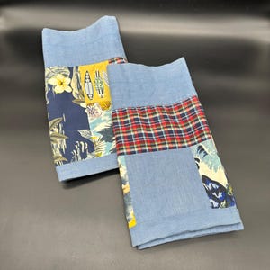 Könnte beinhalten: Zwei blaue Denim-Patchwork-Küchentücher. Eines der Tücher hat ein marineblaues Blumen- und Surfbrettmuster, während das andere ein rot-grün-gelbes Karomuster hat. Die Handtücher sind gefaltet und auf einer dunklen Oberfläche ausgestellt.