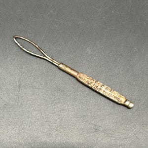 May include: A vintage metal tool with a loop at one end and a textured handle. The handle has engraved text. The tool is silver and gold in color, and is set against a dark gray background.