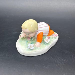 May include: A ceramic figurine of a baby crawling on a patch of grass with flowers. The baby has blonde hair, is wearing a striped shirt, and orange pants. The base is oval-shaped and painted with green and white colors.