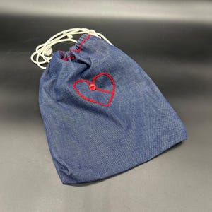 Peut inclure: Un sac en jean bleu avec un cœur rouge brodé et un bouton. Le sac a une fermeture à cordon blanche et des coutures rouges sur le bord supérieur. Le sac est en jean.