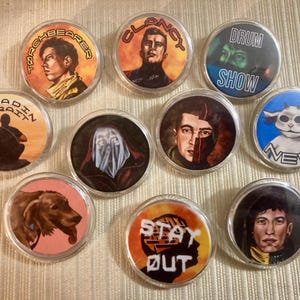Puede incluir: Una colección de diez pines redondos con ilustraciones coloridas. Los diseños incluyen retratos, un perro y textos como "Torchbearer", "Clancy", "Drum Show", "Paladin Strait", "Neo" y "Stay Out".