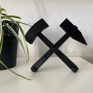 Könnte beinhalten: Eine schwarze 3D-gedruckte Skulptur aus zwei gekreuzten Spitzhacken. Die Spitzhacken bestehen aus glattem, mattschwarzem Kunststoff.