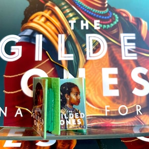 Puede incluir: Una versión en miniatura del libro "The Gilded Ones" de Namwali Serpell. La portada del libro presenta a una mujer de piel oscura y cabello oscuro que lleva un collar de oro y una prenda verde y dorada.