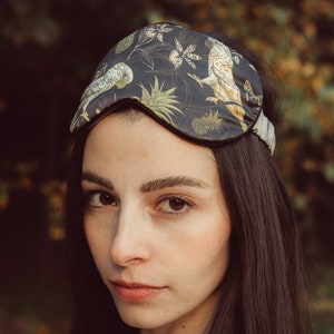 Puede incluir: Una mujer con el pelo largo y oscuro lleva una máscara de dormir negra con estampado floral y de pájaros. La máscara tiene una banda elástica blanca.