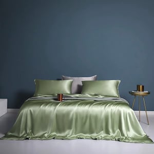May include: A bed dressed with green silk bedding, including a fitted sheet, two pillowcases, and a blanket. Two grey pillows and a brown mug are on the bed. A small gold side table with a gold container is to the right.