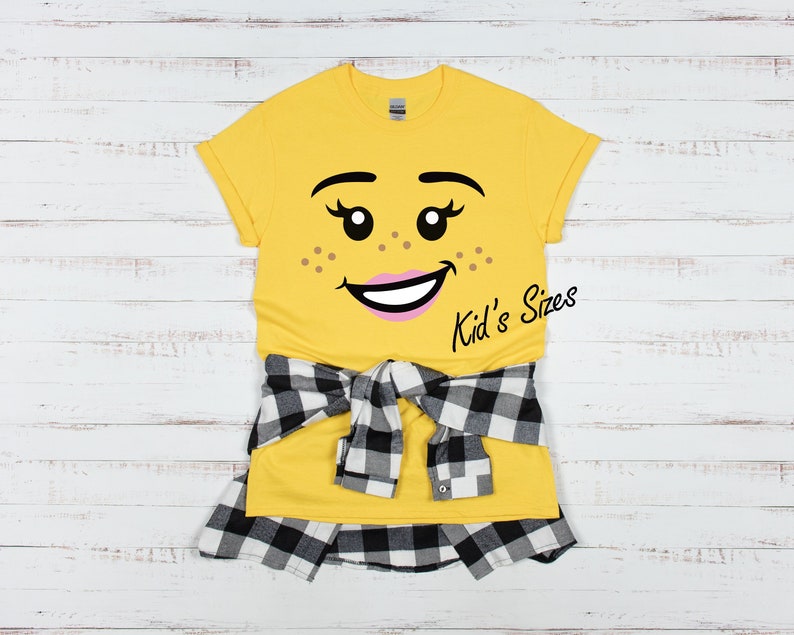 May include: Yellow t-shirt with a cartoon face graphic, freckles, and the text "Kid's Sizes." A black and white plaid shirt is tied around the waist. The shirt is on a white wooden background.