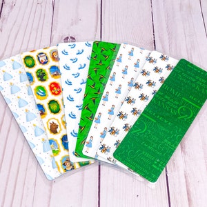 May include: Set of eight bookmarks featuring a Wizard of Oz theme. The bookmarks are made of cardstock and feature various designs, including a green background with a witch's hat, a blue background with ruby slippers, and a white background with a yellow brick road. The bookmarks also feature text from the book, such as "There's no place like home" and "Believe".