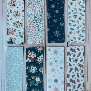 May include: Eight different bookmark designs featuring wintery patterns. The designs include snowflakes, pine cones, floral patterns, and swirls. The bookmarks are printed on white cardstock and have a clear plastic coating.