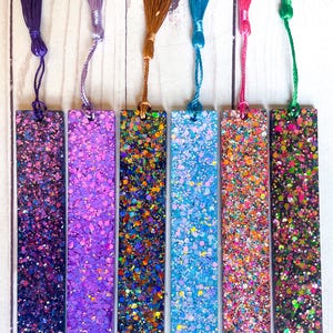 May include: A collection of handmade glitter bookmarks in various colors. Each bookmark features a rectangular shape covered in sparkling glitter and a matching tassel. Colors include purple, brown, blue, pink, and green.