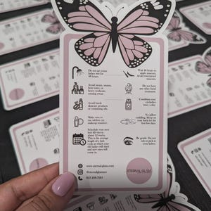 K&ouml;nnte beinhalten: Eine rosa-schwarze Karte in Schmetterlingsform mit Wimpernpflegeanweisungen. Die Karte enth&auml;lt Text und Symbole mit Pflegehinweisen, wie z. B. das Vermeiden von Wasser und Dampf. Der Markenname "Eternal Glam" ist ebenfalls sichtbar.