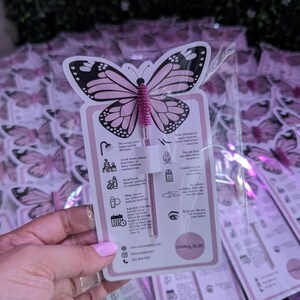 K&ouml;nnte beinhalten: Eine rosa und schwarz geformte Karte in Schmetterlingsform mit einer rosa Mascarab&uuml;rste. Die Karte enth&auml;lt Anweisungen und den Text "Eternal Glam". Der Hintergrund zeigt mehrere &auml;hnliche Karten in Klarsichtverpackungen.