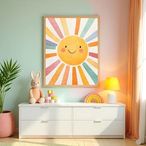 Puede incluir: Un cuadro enmarcado con una acuarela de un sol sonriente con rayos coloridos cuelga sobre una cómoda blanca. Un conejo de peluche, bloques de madera y una decoración en forma de arcoíris están sobre la cómoda. Una lámpara amarilla y una planta en maceta completan la escena.