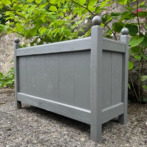 May include: A grey wooden planter box with decorative ball finials on each corner. The planter is sitting on a grey gravel surface.