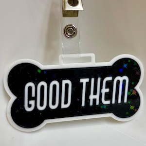 May include: A white and black dog bone-shaped tag with the words "GOOD THEM" in white. The black portion has a holographic, star-like design. A clear plastic clip is attached to the top.