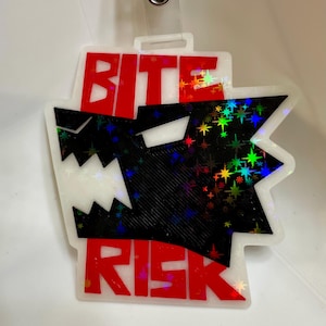 May include: A white, rectangular tag with the words "BITE" and "RISK" in red block letters. A black wolf head graphic with holographic starbursts is in the center. The tag has a clear plastic loop at the top for attachment.