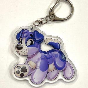 May include: A purple and white inflatable dog keychain with a silver metal ring. The dog is in a playful pose with its tongue sticking out.