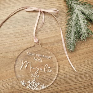 Peut inclure: Boule de Noël transparente en acrylique avec le texte "MON PREMIER NOËL Magalie 2024" et un motif de flocon de neige. Suspendue par un ruban rose, elle repose sur une surface en bois.
