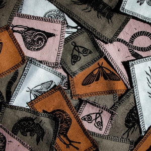 Puede incluir: Una colección de parches de tela con varios diseños inspirados en la naturaleza, incluyendo mariposas, polillas, caracoles y ciempiés. Los parches están cosidos sobre un fondo de tela marrón oscuro.