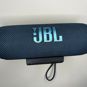 Peut inclure: Un haut-parleur Bluetooth portable JBL bleu avec une sangle noire et un rev&ecirc;tement en tissu textur&eacute;. Le haut-parleur est mont&eacute; sur un mur.