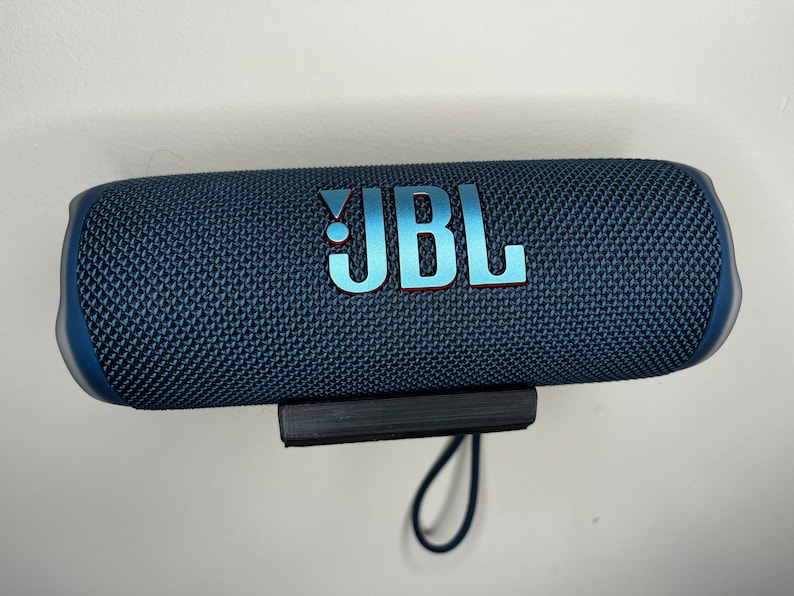 Peut inclure: Un haut-parleur Bluetooth portable JBL bleu avec une sangle noire et un rev&ecirc;tement en tissu textur&eacute;. Le haut-parleur est mont&eacute; sur un mur.