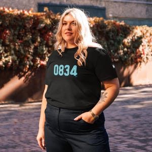 May include: Black t-shirt with the number "0834" in a teal neon font. The shirt also has the letters "AM" in teal below the numbers. The model is wearing dark blue jeans and a gold watch.