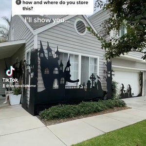 May include: A black metal silhouette of a haunted house, trees, and a graveyard decorates the front of a house for Halloween. The silhouette is attached to the house with screws. The house is painted gray with white trim.