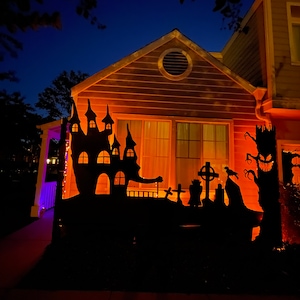 May include: A house decorated for Halloween with a black silhouette of a haunted house, a graveyard, and a spooky tree with a face. The house is lit up with orange lights.