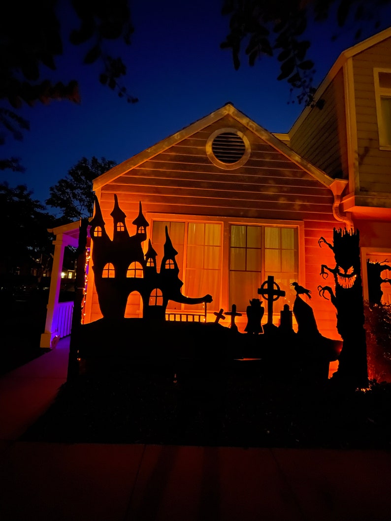 May include: A house decorated for Halloween with a black silhouette of a haunted house, a graveyard, and a spooky tree with a face. The house is lit up with orange lights.