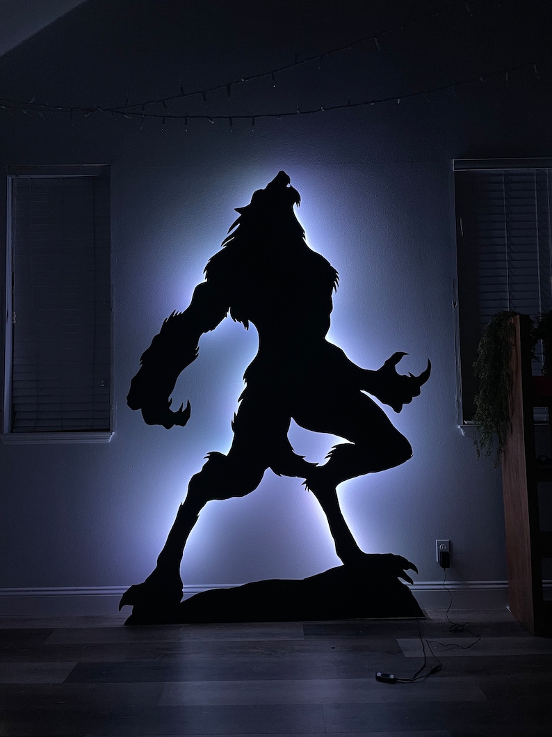 May include: A black silhouette of a werewolf howling at the moon, illuminated with a white light. The silhouette is mounted on a wall and is lit from behind.